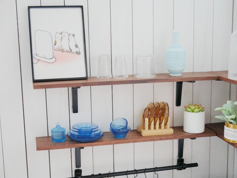 May include: Two wooden shelves with black metal brackets, holding various items including a framed illustration of three cats watching a television, two clear glass cups, a blue and white striped vase, a blue glass jar, a set of blue glass plates, a blue glass bowl, a wooden coaster with six brown glass coasters, a small white planter with a green succulent, and a larger white planter with a green succulent.