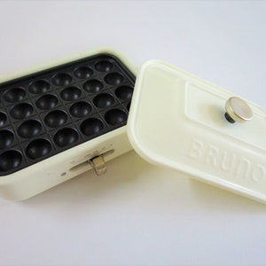 May include: A cream-colored rectangular electric takoyaki pan with a black non-stick cooking surface featuring twenty-four circular indentations. The matching cream lid has the word BRUNO embossed on the front and a metallic knob handle.