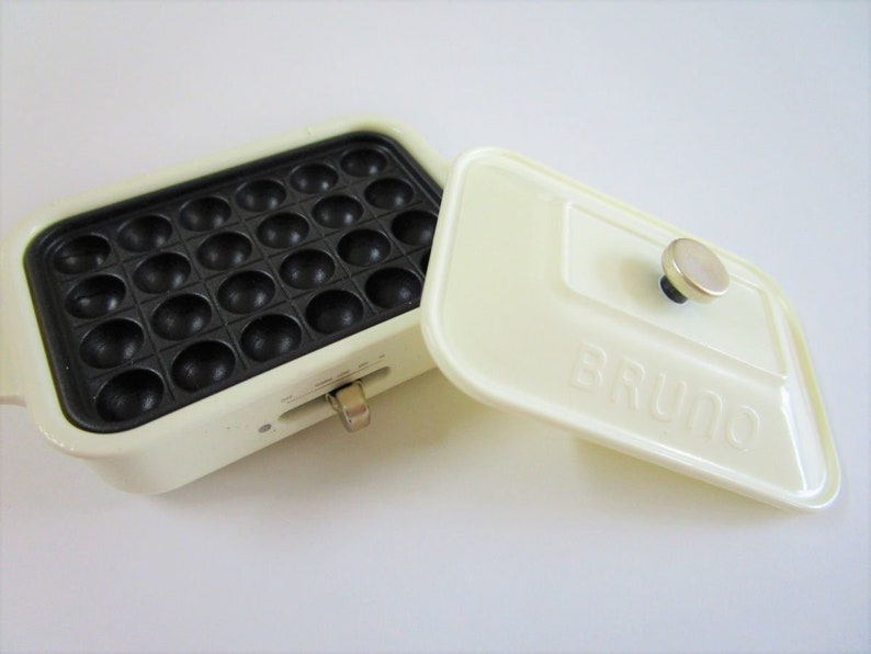 May include: A cream-colored rectangular electric takoyaki pan with a black non-stick cooking surface featuring twenty-four circular indentations. The matching cream lid has the word BRUNO embossed on the front and a metallic knob handle.