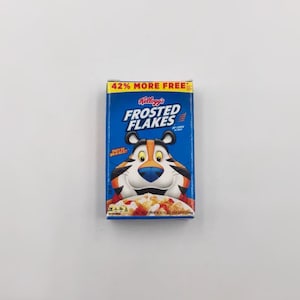 May include: A blue box of Kellogg's Frosted Flakes cereal featuring Tony the Tiger. The box has the text "42% MORE FREE" at the top and "THEY'RE GR-R-REAT!" on the side. The cereal box is on a white background.