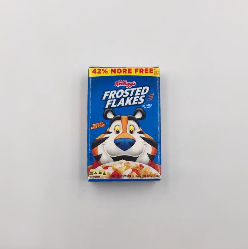 May include: A blue box of Kellogg's Frosted Flakes cereal featuring Tony the Tiger. The box has the text "42% MORE FREE" at the top and "THEY'RE GR-R-REAT!" on the side. The cereal box is on a white background.