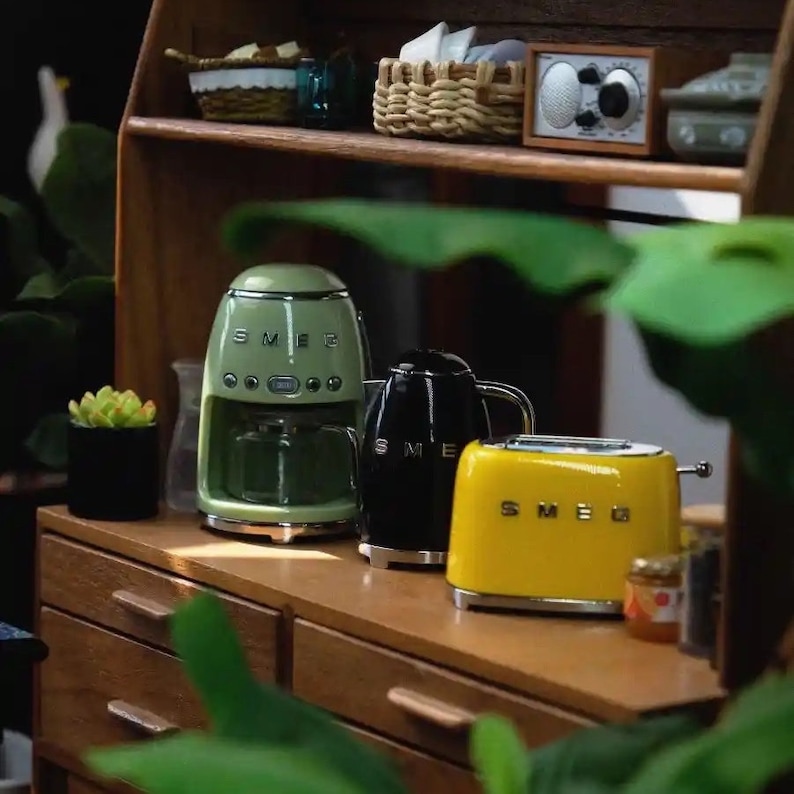 Peut inclure: Ensemble d'appareils de cuisine SMEG de style r&eacute;tro. L'ensemble comprend une cafeti&egrave;re vert clair, une bouilloire noire et un grille-pain jaune. Les appareils sont dispos&eacute;s sur une surface en bois, avec une petite plante succulente et d'autres objets d&eacute;coratifs.