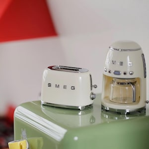 Peut inclure: Ensemble de grille-pain et cafeti&egrave;re SMEG cr&egrave;me. Le grille-pain a deux fentes avec un dessus chrom&eacute; et le logo SMEG. La cafeti&egrave;re a une carafe en verre et des accents chrom&eacute;s. Les deux appareils sont pos&eacute;s sur une surface verte.