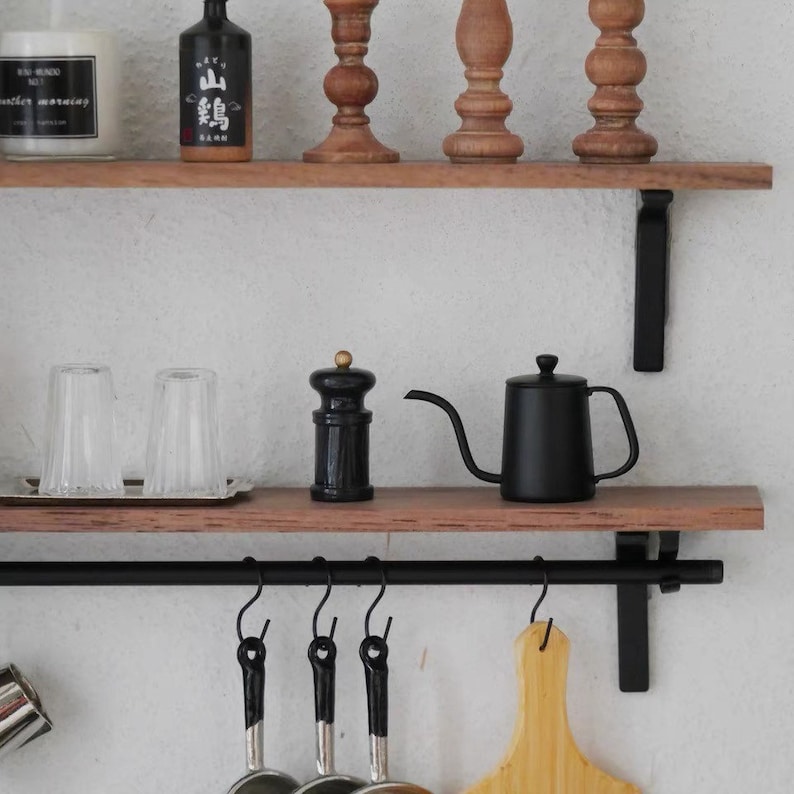 May include: A black metal shelf with two wooden shelves. The top shelf has a bottle of sake, two candles, and three wooden decorative pieces. The bottom shelf has two clear glass cups, a black pepper grinder, and a black metal pour-over coffee pot. The shelf is mounted to a black metal bar with hooks.