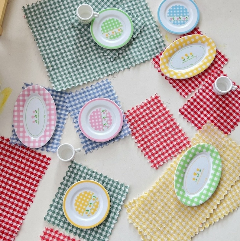 Op de afbeelding: Een set van acht miniatuur borden en kopjes met bloemenmotieven op verschillende geruite placemats. De borden en kopjes zijn wit met roze, gele en blauwe bloemenmotieven. De placemats zijn rood-wit, blauw-wit en groen-wit.