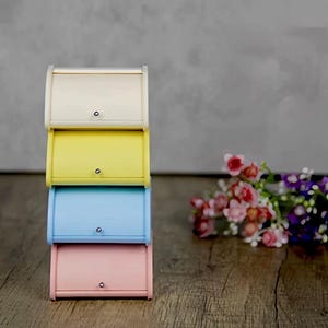 以下が含まれることがあります： クリーム、イエロー、ライトブルー、ピンクの4色の積み重ね可能なパン箱。各箱には、前面に小さな銀色のノブが付いています。