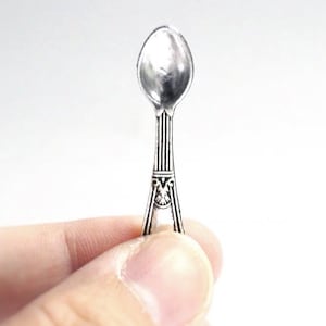 Puede incluir: Una pequeña cuchara plateada con un cuenco ovalado y un mango detallado. El mango presenta líneas verticales y un emblema decorativo cerca del cuenco. La cuchara se sostiene en la mano de una persona sobre un fondo blanco.