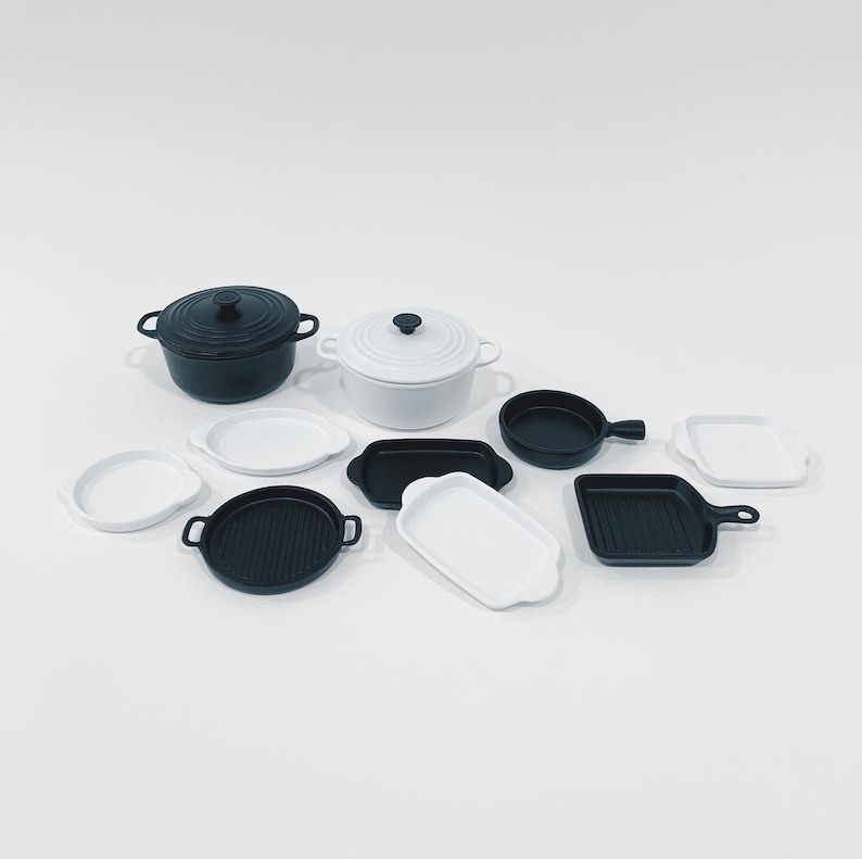 May include: An assortment of miniature cookware in black and white. The collection includes a black Dutch oven, a white Dutch oven, small frying pans, grill pans, and serving dishes. The items are arranged on a white surface.