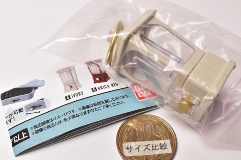 May include: A white and clear plastic miniature model of a vintage music box with a gold-colored top. The model is packaged in a clear plastic bag with a printed instruction sheet. The instruction sheet shows the model in different colors, including ivory and brick red. The sheet also includes a size comparison image with a Japanese 100 yen coin.