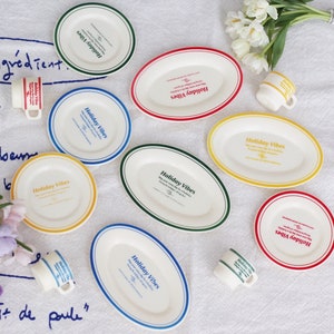 Op de afbeelding: Een set van acht witte ovale borden met kleurrijke randen en de tekst "Holiday Vibes" erop gedrukt. De borden zijn in een cirkel op een wit tafelkleed gerangschikt met een paar witte bloemen en een paar kleine kopjes.