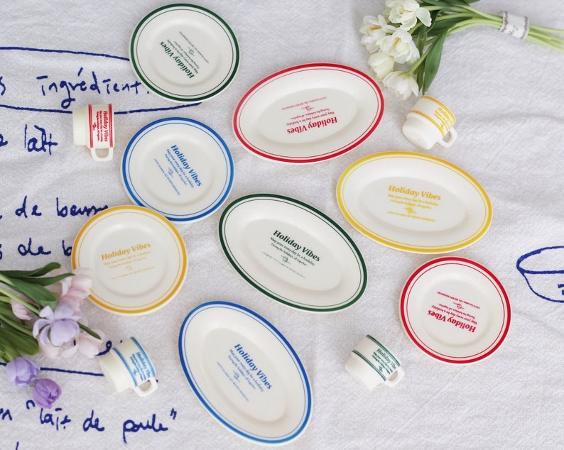 Op de afbeelding: Een set van acht witte ovale borden met kleurrijke randen en de tekst "Holiday Vibes" erop gedrukt. De borden zijn in een cirkel op een wit tafelkleed gerangschikt met een paar witte bloemen en een paar kleine kopjes.