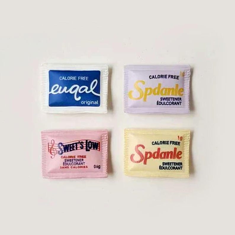 Pode incluir: Quatro pequenos e coloridos pacotes de ado&ccedil;ante sem calorias. Os pacotes s&atilde;o brancos, lavanda, rosa e amarelo, cada um com um nome de marca e design diferentes. As marcas incluem Equal, Spdanle e Sweet's Low. Os pacotes est&atilde;o dispostos em uma superf&iacute;cie branca.