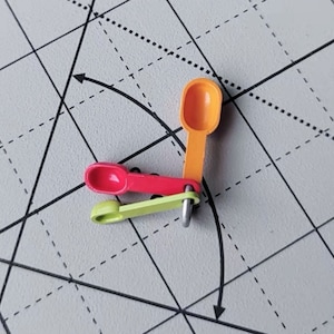 Può includere: Tre cucchiaini in miniatura, uno arancione, uno rosso e uno verde, sono collegati tra loro da un anello di metallo. I cucchiaini sono disposti a forma di triangolo.