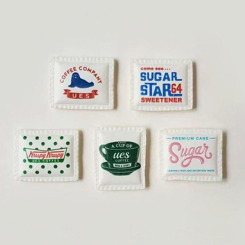 Pode incluir: Uma cole&ccedil;&atilde;o de cinco pequenos pacotes quadrados com v&aacute;rios designs e texto. Os pacotes incluem logotipos de "Coffee Company", "Sugar Star64", "Krispy Kruspy UES Coffee", "A Cup of UES Coffee" e "Sugar". Os pacotes s&atilde;o brancos com gr&aacute;ficos coloridos.
