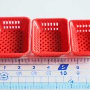 Op de afbeelding: Drie rode plastic manden van verschillende groottes, met een rasterpatroon, zijn uitgelijnd op een witte liniaal met zwarte markeringen. De liniaal meet in centimeters en inches.