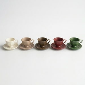 May include: Five ceramic teacups and saucers in a row, each a different color: cream, tan, brown, red, and green. The cups have a classic design with a rounded body and a small handle. The saucers are round and have a matching color.