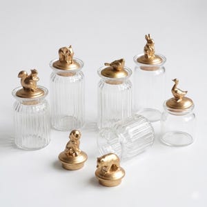 Peut inclure: Ensemble de bocaux en verre transparent avec des côtés cannelés, chacun surmonté d'une figurine d'animal dorée. Les figurines représentent un écureuil, un éléphant, un oiseau, un lapin, un chien, un ours et un canard. Sur fond blanc.