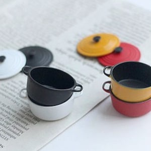 May include: Miniature colorful cookware set with lids. The set includes a white, black, yellow, and red pot. The lids are black, white, yellow, and red. The pots have small handles. The cookware is on a newspaper.