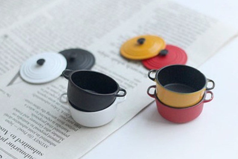 May include: Miniature colorful cookware set with lids. The set includes a white, black, yellow, and red pot. The lids are black, white, yellow, and red. The pots have small handles. The cookware is on a newspaper.