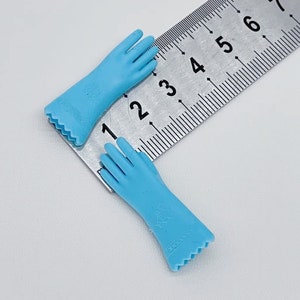 May include: Two light blue doll gloves with detailed fingers and a decorative cuff. The gloves are approximately 2 inches long. The gloves are made of a smooth material and are positioned on a metal ruler.