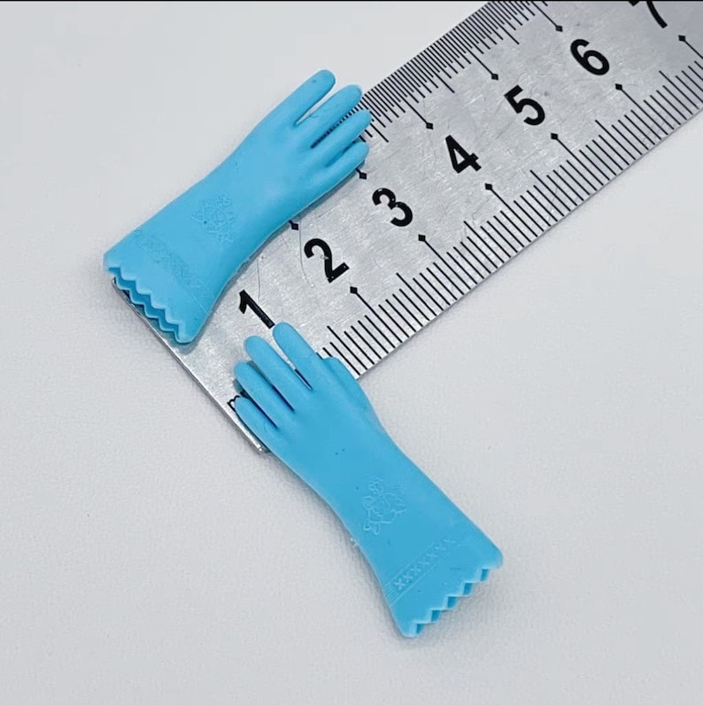 May include: Two light blue doll gloves with detailed fingers and a decorative cuff. The gloves are approximately 2 inches long. The gloves are made of a smooth material and are positioned on a metal ruler.