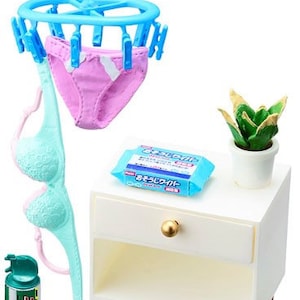 Op de afbeelding: Een wit nachtkastje met een lade en een klein plantje erop. Er hangt een blauwe en roze lingerie set aan een blauwe kledingrek, een blauwe en witte bh en een blauw en wit pakketje op het nachtkastje.