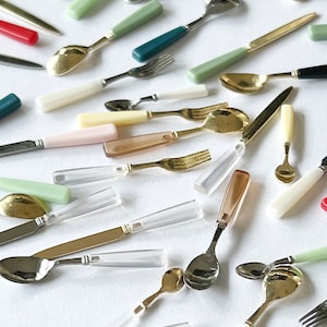May include: A collection of miniature spoons and forks with various colored handles, including green, blue, yellow, pink, and white. The handles are made of plastic and the utensils are made of metal.