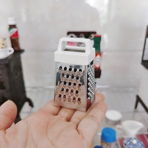 Op de afbeelding: Een kleine, witte en zilveren rasp met een handvat. De rasp heeft een rechthoekige vorm en is gemaakt van metaal. Het is een keukengereedschap dat wordt gebruikt om kaas, groenten en andere voedingsmiddelen te raspen.