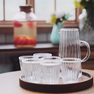 May include: A set of six clear glass drinking glasses with a ribbed texture, and a clear glass jug with a handle, all on a wooden tray.