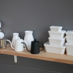 May include: A white shelf with a variety of white kitchenware, including a teapot, a pitcher, and a vase. There are also several white plastic containers stacked on the shelf.