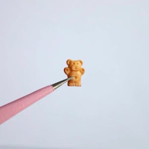 May include: A small, brown bear-shaped cookie is held by a pair of pink tweezers against a white background.
