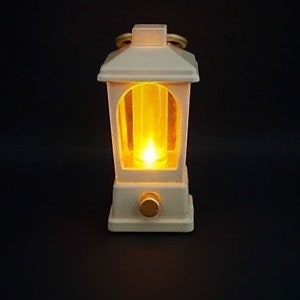 May include: A white lantern with a warm yellow light shining through the glass panels. The lantern has a decorative handle and a small knob on the bottom.
