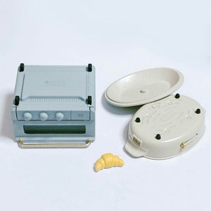 May include: A miniature light blue toaster oven with a silver handle and three knobs, an oval-shaped beige lidded dish with four small black feet, and a single small yellow croissant figurine. These items are designed as small-scale kitchen accessories.