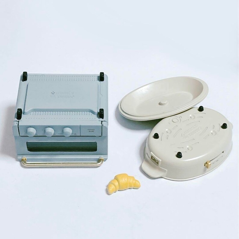 May include: A miniature light blue toaster oven with a silver handle and three knobs, an oval-shaped beige lidded dish with four small black feet, and a single small yellow croissant figurine. These items are designed as small-scale kitchen accessories.