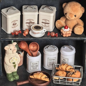 Può includere: Una scena di cucina in miniatura con barattoli bianchi con le etichette "tea", "coffee" e "sugar". Sono presenti anche un orsacchiotto, pasticcini e una statuina di maiale. La scena è ambientata su una mensola scura, creando un'atmosfera accogliente e invitante.