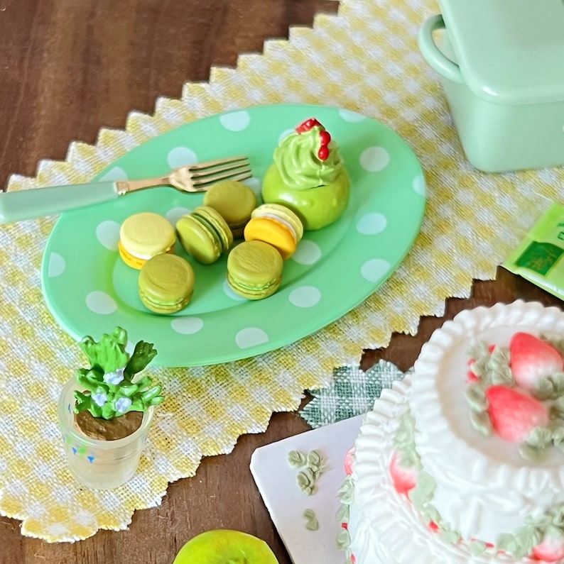 Op de afbeelding: Een groen en wit gestippeld bord met verschillende groene en gele macarons en een groene en witte cupcake met rode sprinkles. Het bord ligt op een geel en wit geruit tafelkleed.