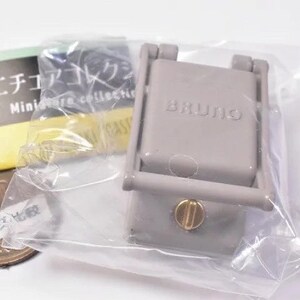 May include: A miniature grey BRUNO kitchen appliance replica encased in clear plastic packaging. The device features a rectangular top with the brand name embossed, a hinged frame, and a small gold-colored circular knob on the front panel.