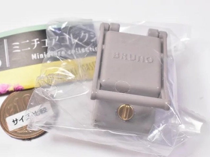 May include: A miniature grey BRUNO kitchen appliance replica encased in clear plastic packaging. The device features a rectangular top with the brand name embossed, a hinged frame, and a small gold-colored circular knob on the front panel.