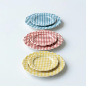 May include: Three sets of two stacked ceramic plates, each featuring a light blue, pink, or yellow gingham check pattern. The plates have scalloped edges and a glossy finish, arranged vertically in a column against a plain white background.