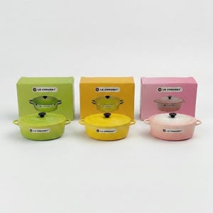 Puede incluir: Tres pequeñas cocottes Le Creuset de hierro fundido de colores verde lima, amarillo y rosa, cada una con una tapa a juego y un pomo negro. Cada cocotte se muestra frente a su caja a juego.