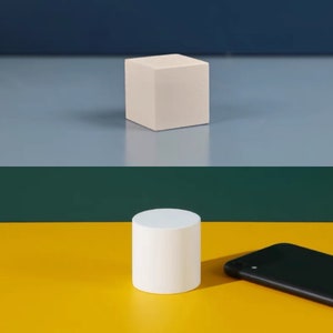 Puede incluir: Un cubo blanco y un cilindro blanco sobre una superficie amarilla. Un teléfono inteligente negro también está sobre la superficie.