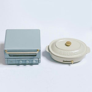 May include: A light blue toaster oven with a gold handle and three control knobs, positioned next to an oval-shaped cream-colored electric griddle with a gold lid knob and the word BRUNO embossed on the front of the lid.