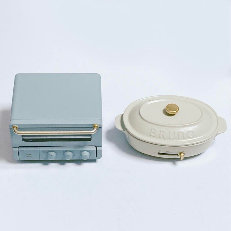 May include: A light blue toaster oven with a gold handle and three control knobs, positioned next to an oval-shaped cream-colored electric griddle with a gold lid knob and the word BRUNO embossed on the front of the lid.