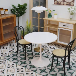 May include: A white round table with a white pedestal base sits in a room with a black and white tiled floor. Two black chairs with woven cane seats are placed around the table. A gold floor lamp with a white shade is in the background.