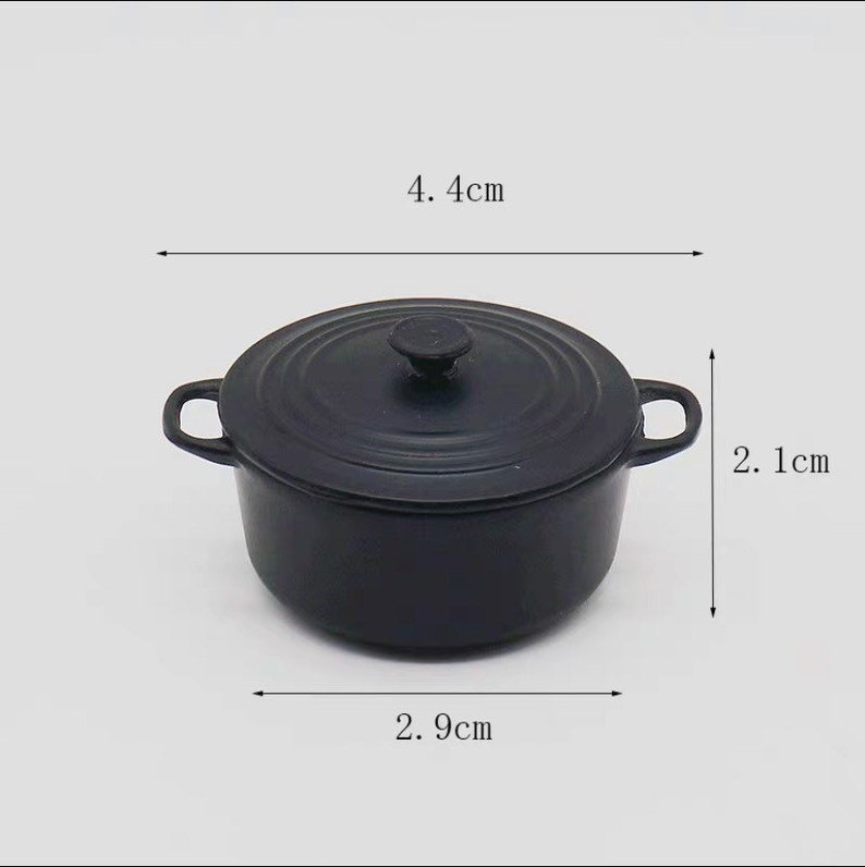 May include: A small, black cast iron pot with a lid and two handles. The pot measures 4.4 cm in diameter, 2.9 cm wide, and 2.1 cm tall. The pot is designed for cooking and is suitable for a miniature kitchen.