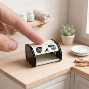 Puede incluir: Miniatura de panera con cara de panda, con ribete negro y tapa abatible. La cara del panda está pintada en la parte delantera, con ojos negros y una pequeña sonrisa. La panera está sobre una superficie de madera.