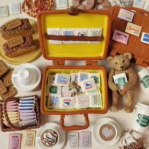 Pode incluir: Uma cena de piquenique em miniatura com um ursinho de pel&uacute;cia, malas cheias de saquinhos de a&ccedil;&uacute;car, x&iacute;caras de caf&eacute; e doces. A cena &eacute; montada em uma superf&iacute;cie branca com v&aacute;rios alimentos.