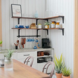 May include: A white kitchen with wooden shelves and a black metal bar hanging from the shelves. The shelves are filled with various kitchen items, including glasses, mugs, plates, and jars. There is a coffee maker on the counter below the shelves. The kitchen has a white countertop and white cabinets. There is a wooden table in the foreground.