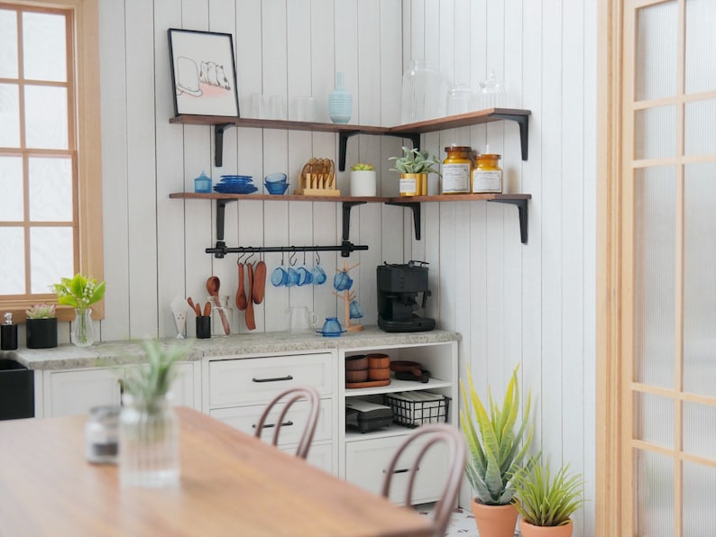 May include: A white kitchen with wooden shelves and a black metal bar hanging from the shelves. The shelves are filled with various kitchen items, including glasses, mugs, plates, and jars. There is a coffee maker on the counter below the shelves. The kitchen has a white countertop and white cabinets. There is a wooden table in the foreground.