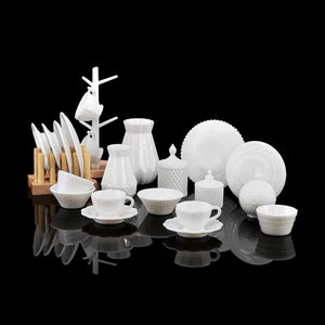 Op de afbeelding: Een verzameling witte melkglas servies, waaronder borden, kommen, kopjes en vazen. Het servies heeft een delicaat, gegraveerd ontwerp en is gerangschikt op een zwart oppervlak.